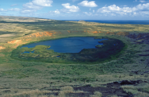 Une vue du cratère du Rano Raraku ; le lac est un lieu de baignade. Sur la pente, on distingue des têtes de moai, émergeant de l’herbe haute.