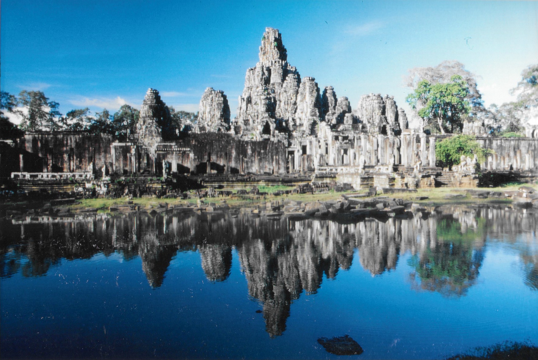 Le somptueux temple du Bayon se mirant dans les eaux de ses douves. Son nom est dérivé d’une appellation signifiant palais du dieu Indra. Dû à Jayavarman VII, pour un culte bouddhique shivaite, il est daté de la toute fin du XIIe siècle et du début du XIIIe. On écrit Bayon, mais on prononce Bayonne, comme la ville française du Pays basque. Il est situé au centre géographique de l’immense enceinte d’Angkor Thom. Le temple est entouré d’une muraille dont les tours affichent quatre visages apparemment sereins, sinon mystérieusement indifférents. Des “Mona Lisa” asiatiques de pierre en quelque sorte....